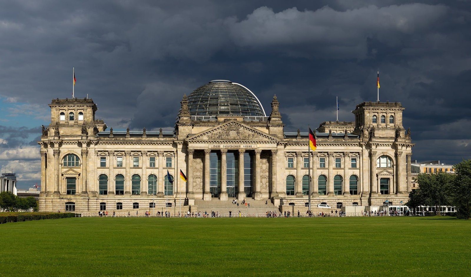 The Reichstag building with its glass dome, sunlit under a dark stormy sky, seen across the lawn to the west